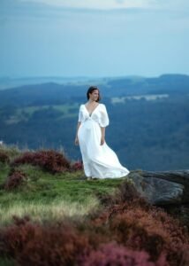 Graceful woman in a flowing white dress stands atop Mother Cap, her presence as ethereal as the surrounding heather.