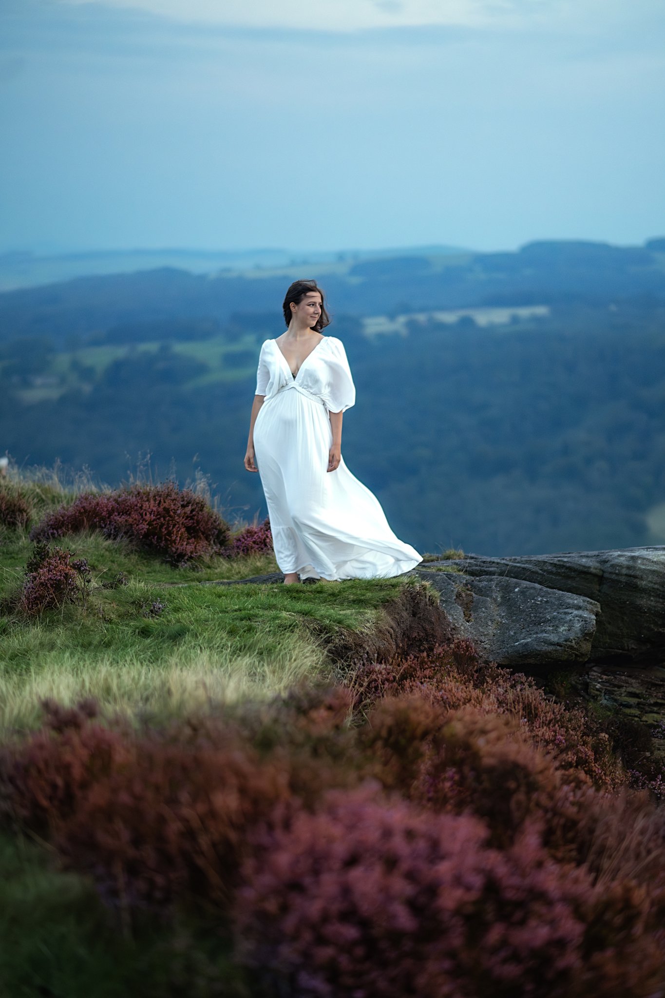Graceful woman in a flowing white dress stands atop Mother Cap, her presence as ethereal as the surrounding heather.