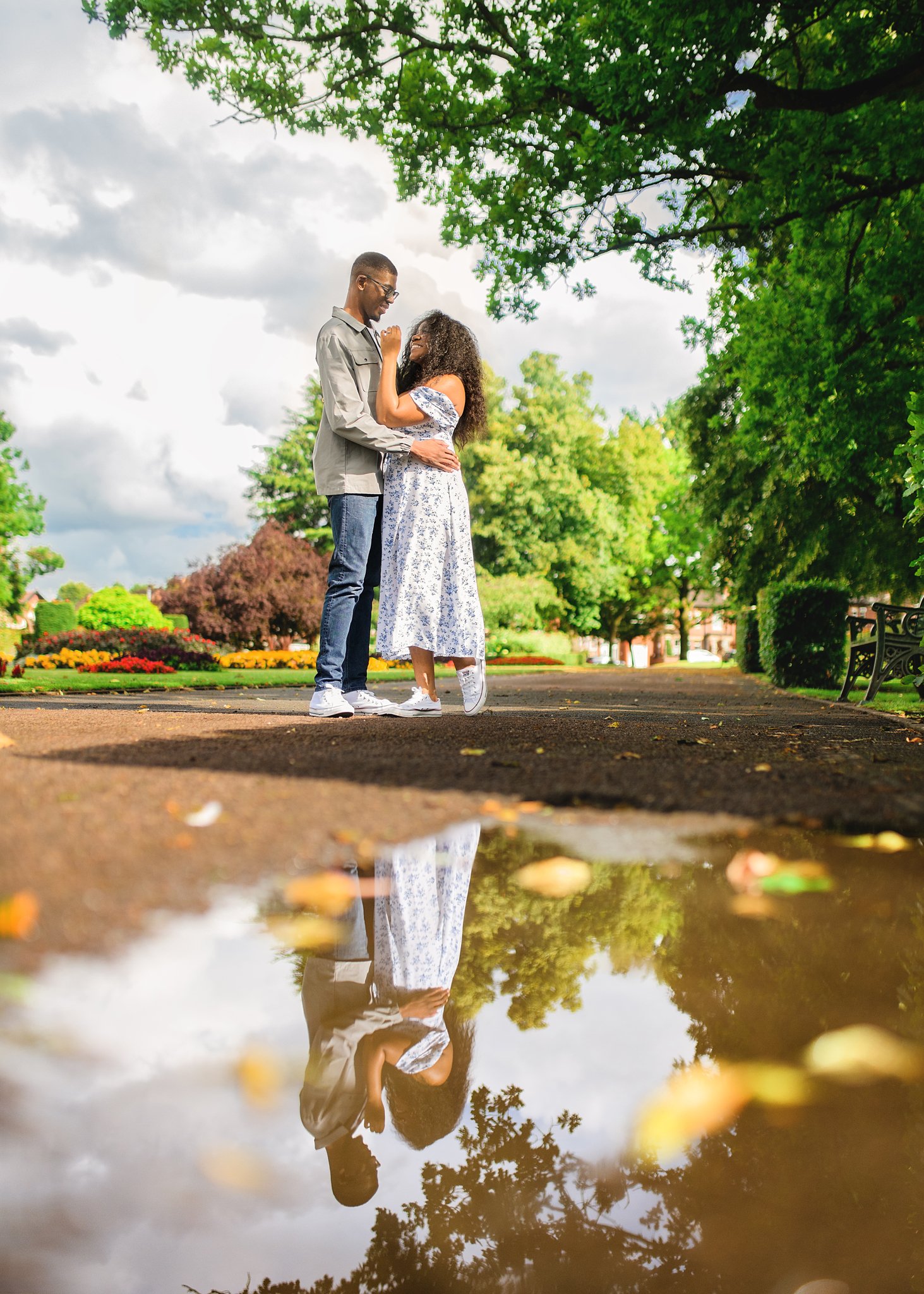 Engaged couple embracing by a reflective water puddle in Nottingham, with the garden's beauty mirrored in the water