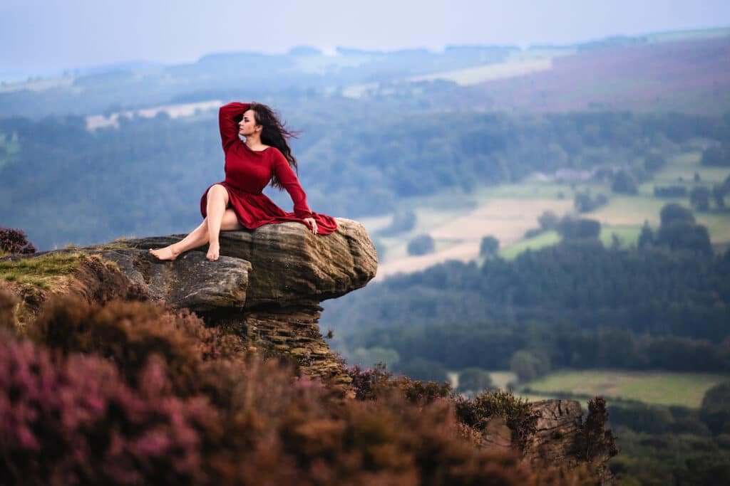 A woman in a red dress seated on a cliff edge, her hair dancing with the wind, overlooking a verdant valley.