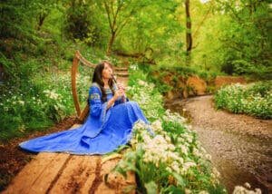A serene harpist in a flowing blue gown sits by a woodland stream, her instrument beside her, enveloped by the lush greenery of a Nottingham forest.