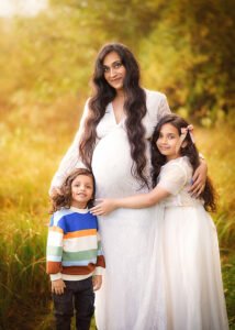 A pregnant mother in a white lace dress stands in a golden meadow with her two children, her daughter in a tulle dress and her son in a colorful sweater, as they await the new family member.