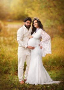 A radiant expectant couple stands embraced in an idyllic natural setting, with the mother-to-be in a white lace gown and floral headpiece symbolizing the purity and fullness of maternity.