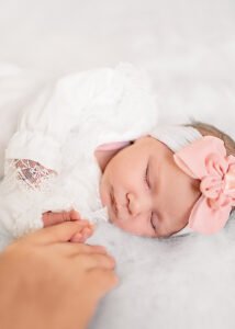 A newborn sleeps soundly, adorned with a pink bow headband, as a loving hand reaches out, in a tender lifestyle newborn photography moment.