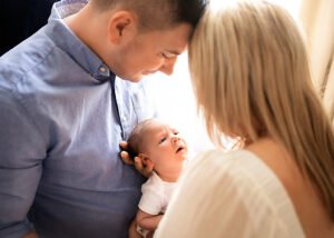 A newborn gazes up at their parents, a moment of early connection captured against the bright backdrop of a window in a lifestyle newborn photograph.