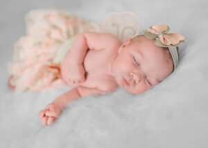 Newborn baby in peaceful sleep adorned with a golden bow headband, lying on a fluffy white backdrop with a soft floral blanket.