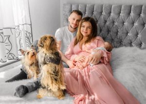 A joyful family portrait with a newborn cradled in the mother's arms, flanked by two attentive dogs, symbolizing a complete family in lifestyle newborn photography.
