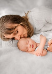Grandmother tenderly kisses her grandchild's forehead as they both lie on a fluffy bed, a serene moment in a lifestyle newborn photography session.