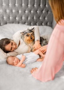 Cozy family moment with a sleeping newborn, a doting grandmother, and an attentive Yorkie on a plush bed, captured in a lifestyle newborn photography setting.