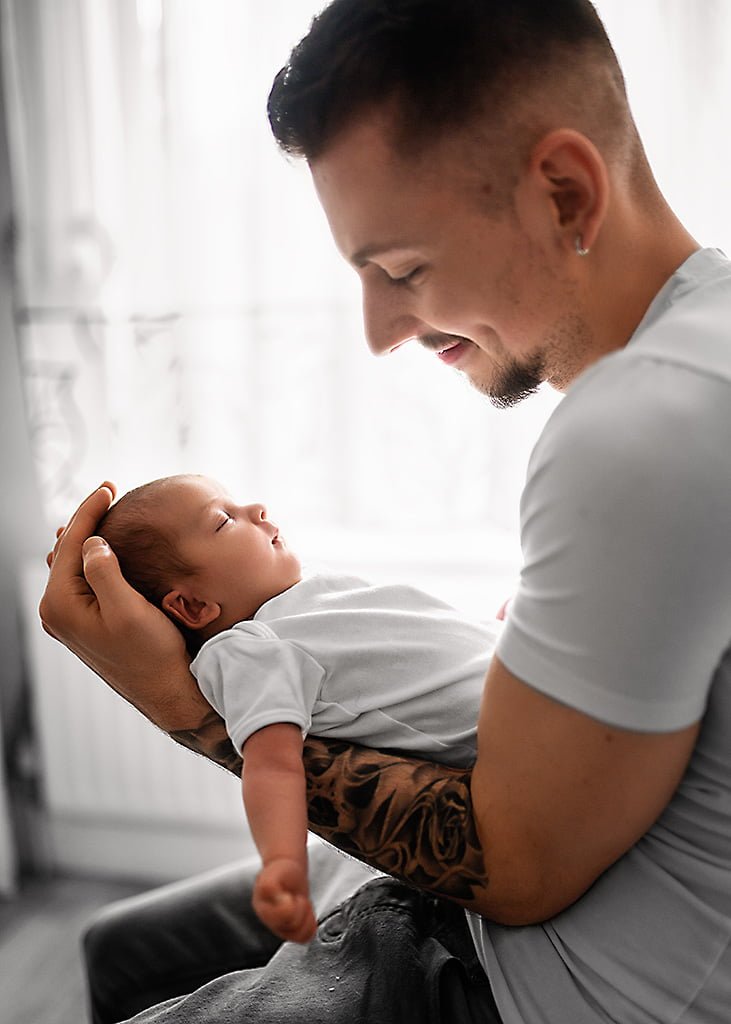 Father holding his newborn baby with a loving smile, as the infant stretches in comfort, captured in a lifestyle newborn photography moment.
