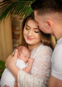 Content newborn sleeps peacefully in mother's arms while father lovingly touches the baby's head, with tropical foliage softening the background in a lifestyle newborn photography portrait.