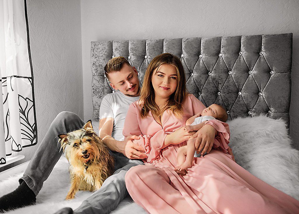New parents and their content newborn are joined by a joyful family dog on a cosy bed, encapsulating a perfect lifestyle newborn photography scene.