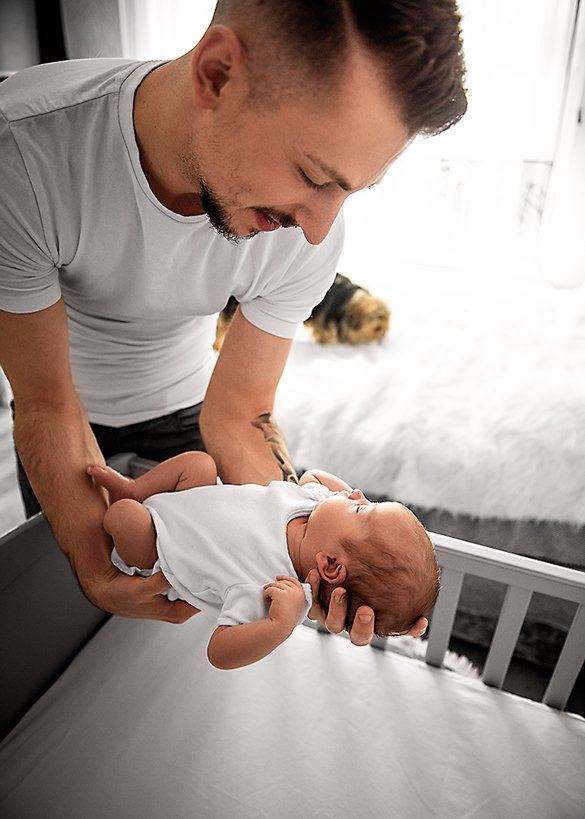 Father cradling his sleeping newborn with gentle hands in a natural light-filled room, showcasing the bond in a lifestyle newborn photography setting.