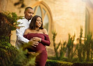 A happy couple in a serene embrace, with the man standing behind the woman, his hands gently wrapped around her burgeoning belly, both dressed elegantly against an old stone building backdrop.