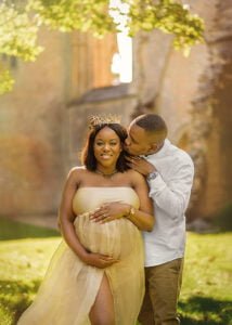 Expectant mother in a flowing golden gown wearing a crown, embraced by her partner during a maternity photoshoot at a historic Nottingham location.