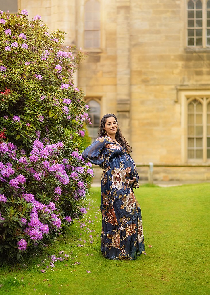 An expectant mother in a floral dress stands beside a vibrant shrub of purple flowers, her hand gently resting on her belly.