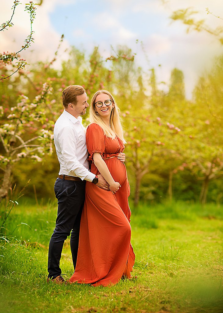 A radiant couple embraces in a lush green field, the pregnant woman in a vibrant orange gown, her joy matched by her partner's affectionate gaze.