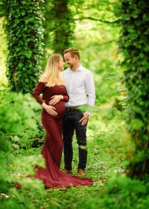Expectant couple in a lush green forest, the mother-to-be wearing a burgundy gown during their maternity photoshoot in Nottingham.