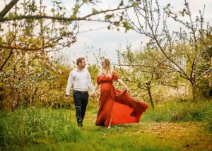 Expectant parents joyfully walking hand in hand through a blossoming orchard during their Nottingham maternity photoshoot, with the mother's dress flowing in the breeze.
