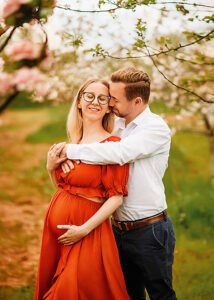 A loving couple in a sunlit orchard, the pregnant woman in a red dress smiling as her partner kisses her forehead, surrounded by delicate pink blossoms.