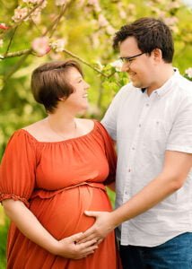 Expectant couple smiling and cradling the baby bump in an orchard during their maternity photoshoot in Nottingham.