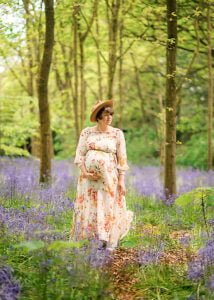 Expectant mother in a floral gown walking through a vibrant bluebell forest during a maternity photoshoot in Nottingham.