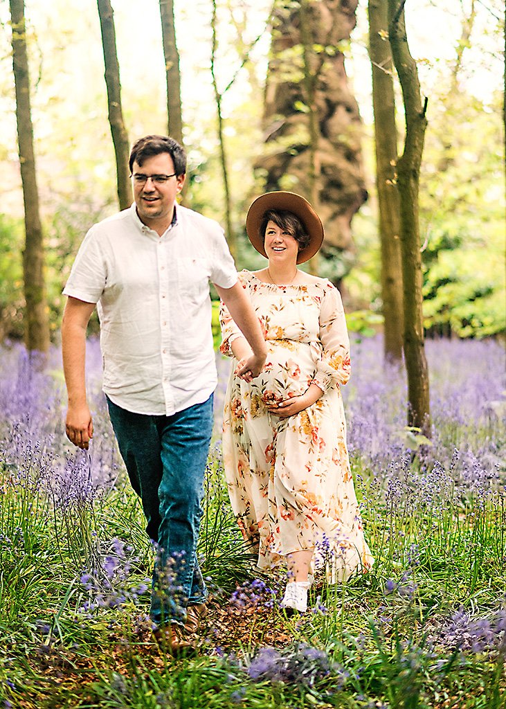 A joyful couple walks hand-in-hand through a purple bluebell forest, with the woman in a floral dress and wide-brimmed hat, showcasing her baby bump.
