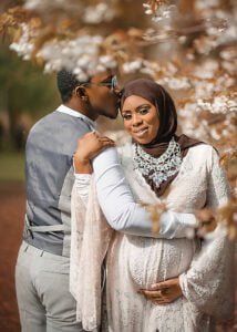 Expectant couple embracing under spring blossoms, with the mother-to-be in a white gown and the father-to-be gently kissing her forehead.