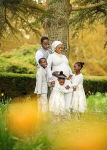 A joyful family embraces around the expectant mother, glowing with happiness in a sunlit Nottingham park during a maternity photoshoot.