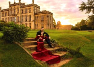A couple enjoys a tender moment on the steps of a grand historic mansion at sunset, with the expectant mother in a stunning red gown that flows elegantly down the stairs.