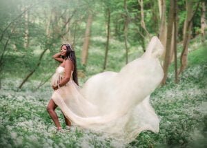A radiant pregnant woman in a flowing dress stands amidst a forest blanketed in white flowers, her happiness as palpable as the spring around her.
