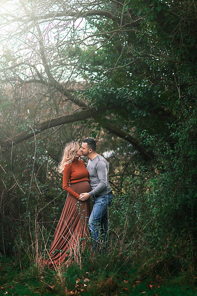 A couple shares a romantic kiss in a secluded Nottingham grove, celebrating their future together in a maternity photoshoot.