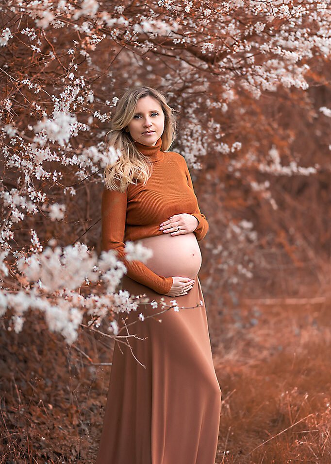 A beautiful expectant mother in earth tones stands among white spring blossoms, her tender touch on her belly conveying the wonder of her journey.