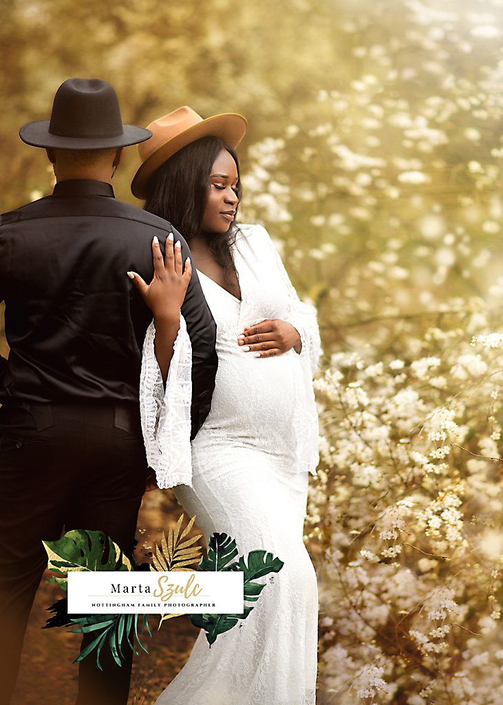 An elegant couple in a maternity photoshoot, with the expectant mother in white and the supportive father by her side, amidst a blossoming Nottingham backdrop.