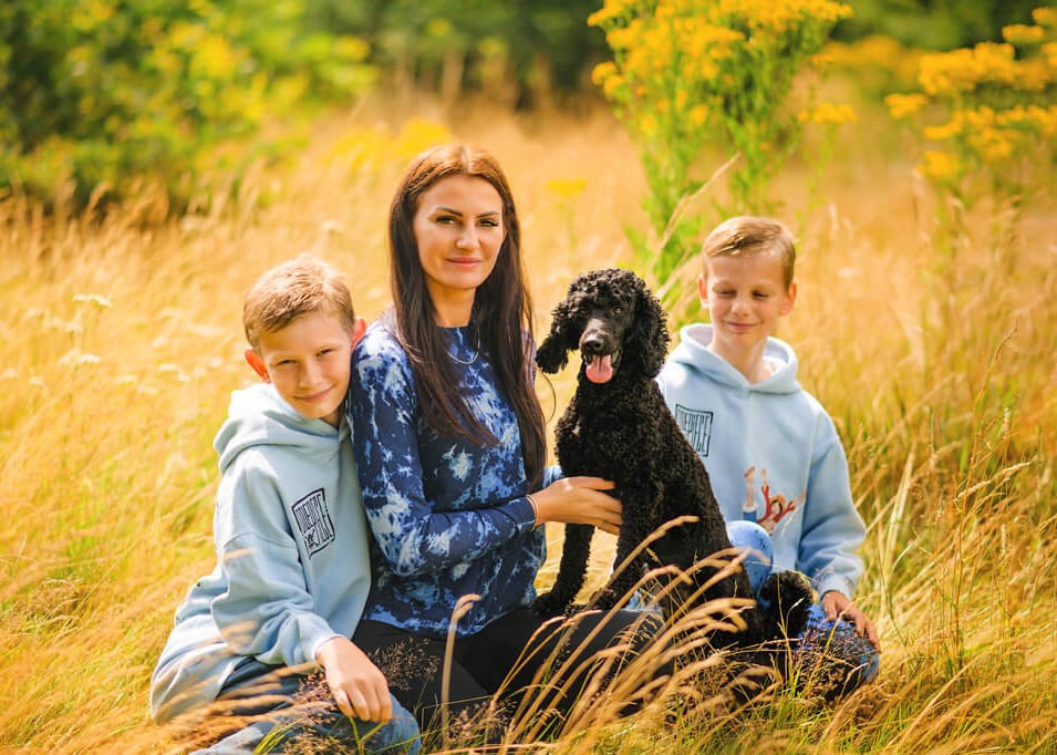 A heartwarming family moment as a smiling woman and two boys cuddle with their black poodle in a golden field, epitomizing family pet photography in Nottingham.