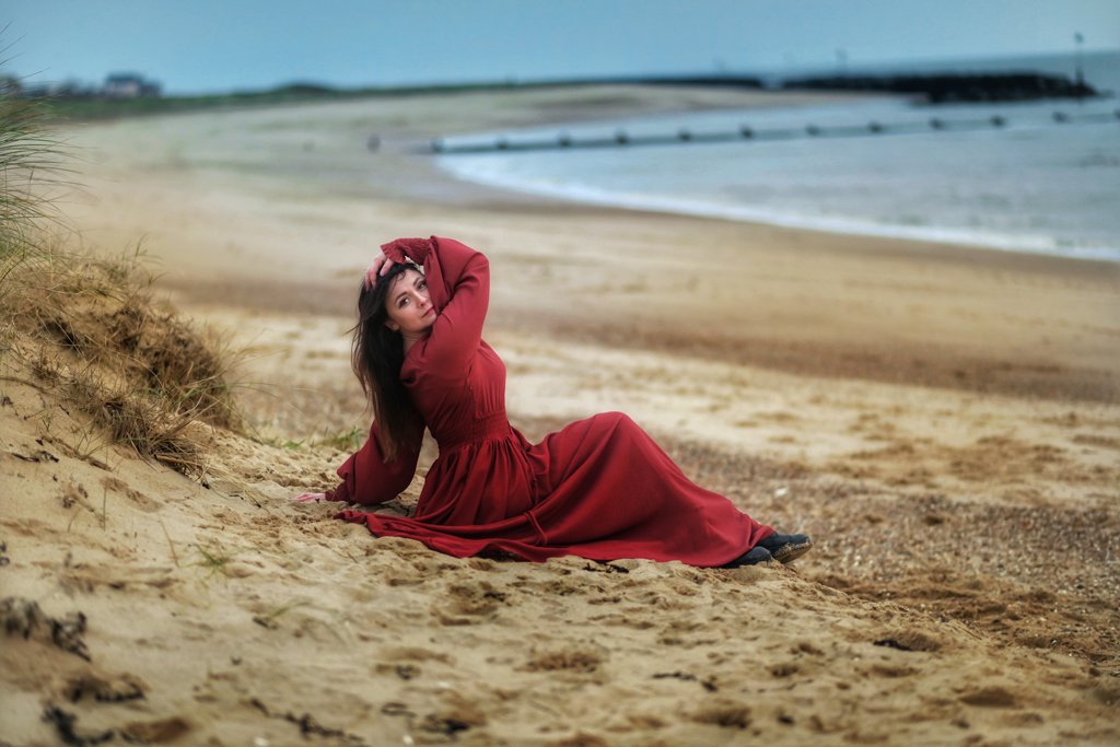 Elegant woman in a flowing red dress reclining on a sandy dune, her hand gracefully touching her head, during a windswept outdoor birthday photoshoot at the beach.