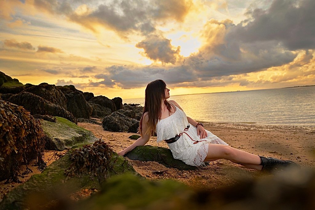 A serene scene of a woman lounging in a delicate white lace dress on a moss-covered rock, with a golden sunset over the calm sea during an outdoor birthday photoshoot.