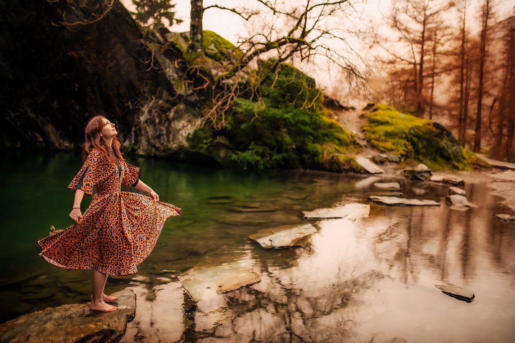 Joyful woman twirling in a leopard-print dress by a serene forest river during an outdoor birthday photoshoot.