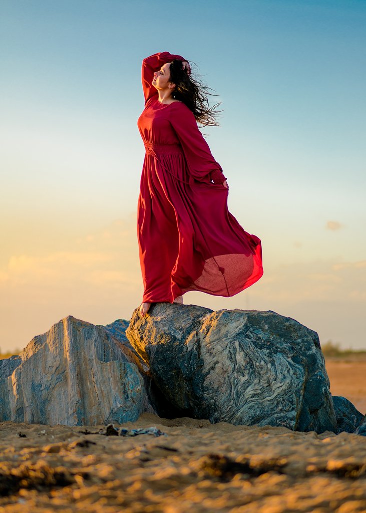 Woman in vibrant red dress stands empowered on a beach rock during an outdoor birthday photoshoot at sunset.