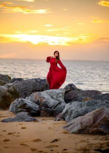 Woman in a stunning red dress stands poised among seaside rocks, her figure framed by a vibrant sunset, in an outdoor birthday photoshoot.