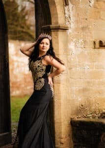 Captivating outdoor birthday photoshoot featuring a woman in an ornate black gown with gold embroidery, crowned with a tiara, next to a historic stone doorway.