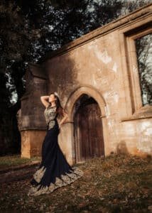 A majestic outdoor birthday photoshoot with a woman in a black and gold gown, radiating regal elegance against an ancient stone archway.