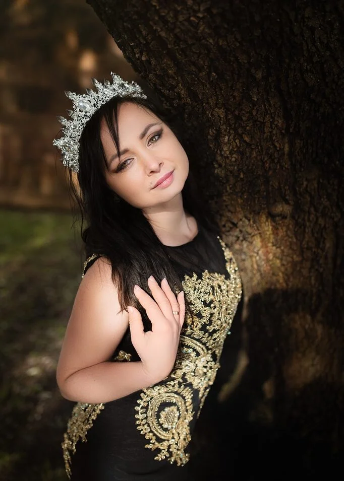 A serene outdoor birthday photoshoot with a woman in a luxurious black gown adorned with intricate gold detail, leaning against an ancient tree.