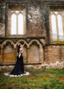 An awe-inspiring outdoor birthday photoshoot captures a woman in a black and gold dress, looking up at the gothic architecture of an ancient ruin.