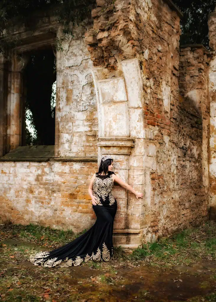 A woman in a black and gold gown with a regal tiara poses against the textured backdrop of ancient stone ruins for an outdoor birthday photoshoot.