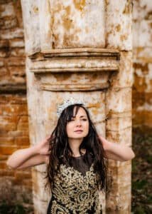 A woman in an ornate black gown with golden embroidery and a sparkling tiara stands against rustic ancient columns in an outdoor birthday photoshoot.