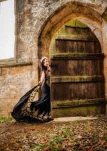 A woman dressed in a black and gold embroidered gown poses beside a vintage wooden door, adding a touch of elegance to an outdoor birthday photoshoot.