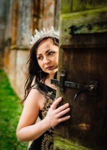A woman adorned with a sparkling tiara peers around a historical wooden door during an outdoor birthday photoshoot.