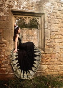 Woman in an ornate gown with golden embroidery posing in a gothic window frame during an outdoor birthday photoshoot.