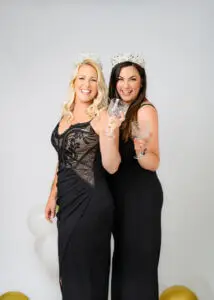 Two women in black dresses smiling and toasting with crystal glasses during a 40th birthday photoshoot.